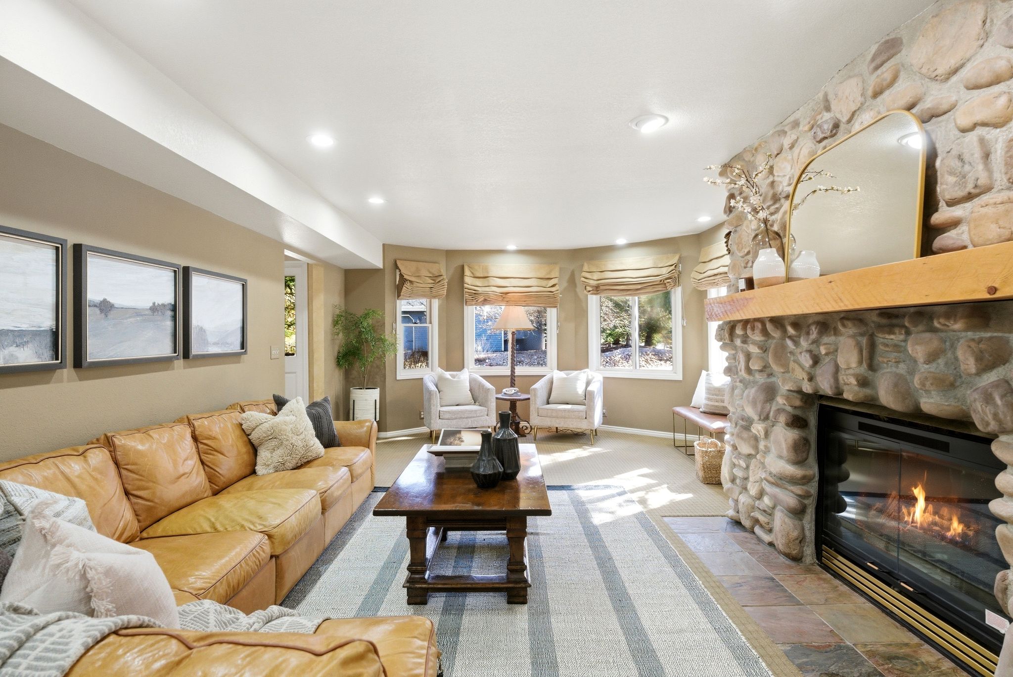 Spacious living area featuring a prominent stone fireplace with a wood mantle, recessed lighting, and multiple windows with Roman shades