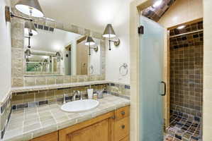 Bathroom featuring a wood-finish vanity with an oval basin, a tile countertop, and a full-width mirror with sconce lighting