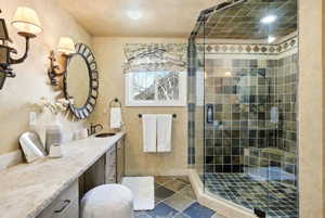 Bathroom featuring a glass-enclosed shower with tile surround and integrated bench seating, a dual-sink vanity with stone countertop, and an oval mirror