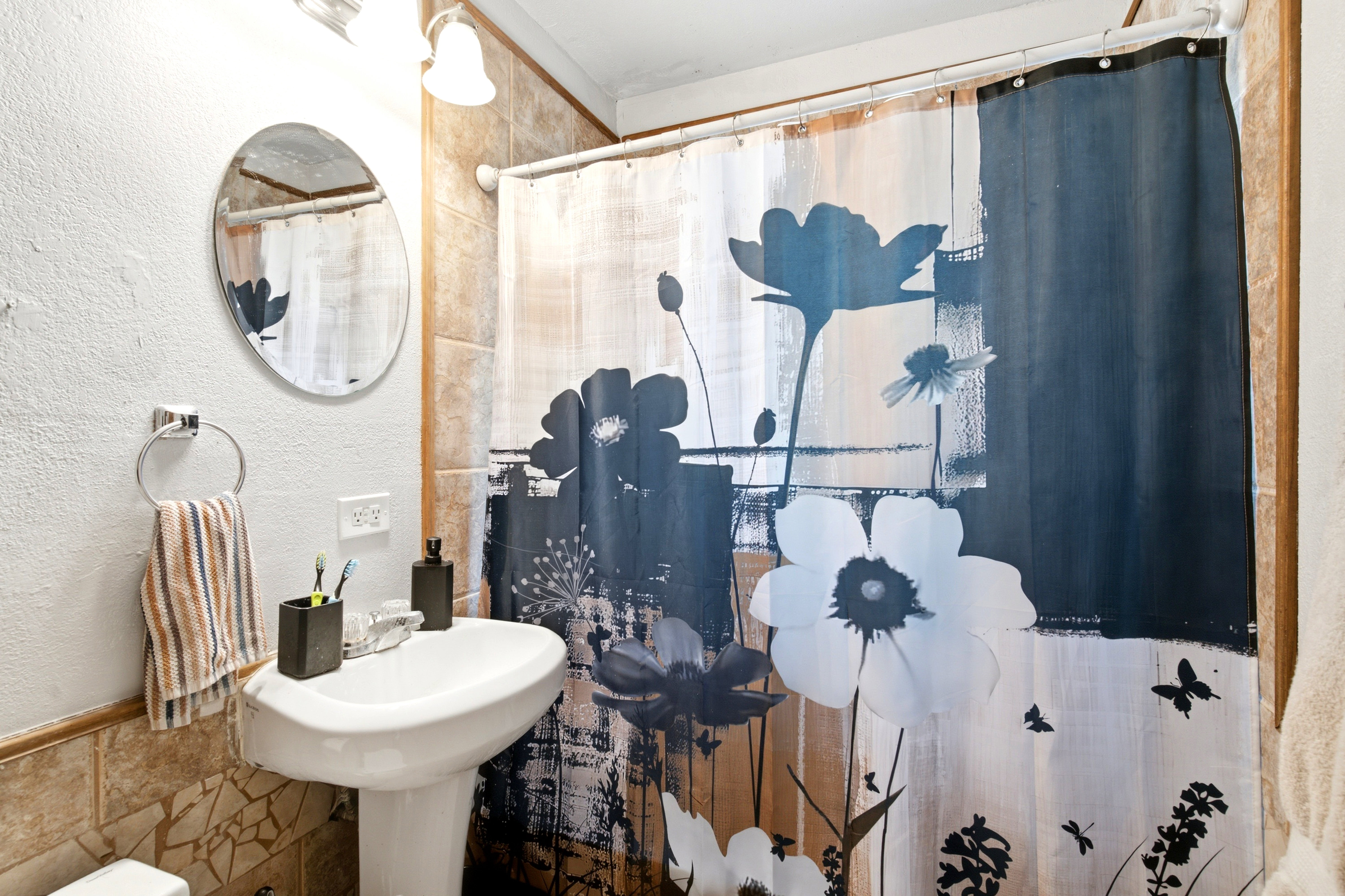 Bathroom featuring a white pedestal sink, an oval wall-mounted mirror, a chrome towel ring, a shower area with a curtain rod, and light-toned wall tiling
