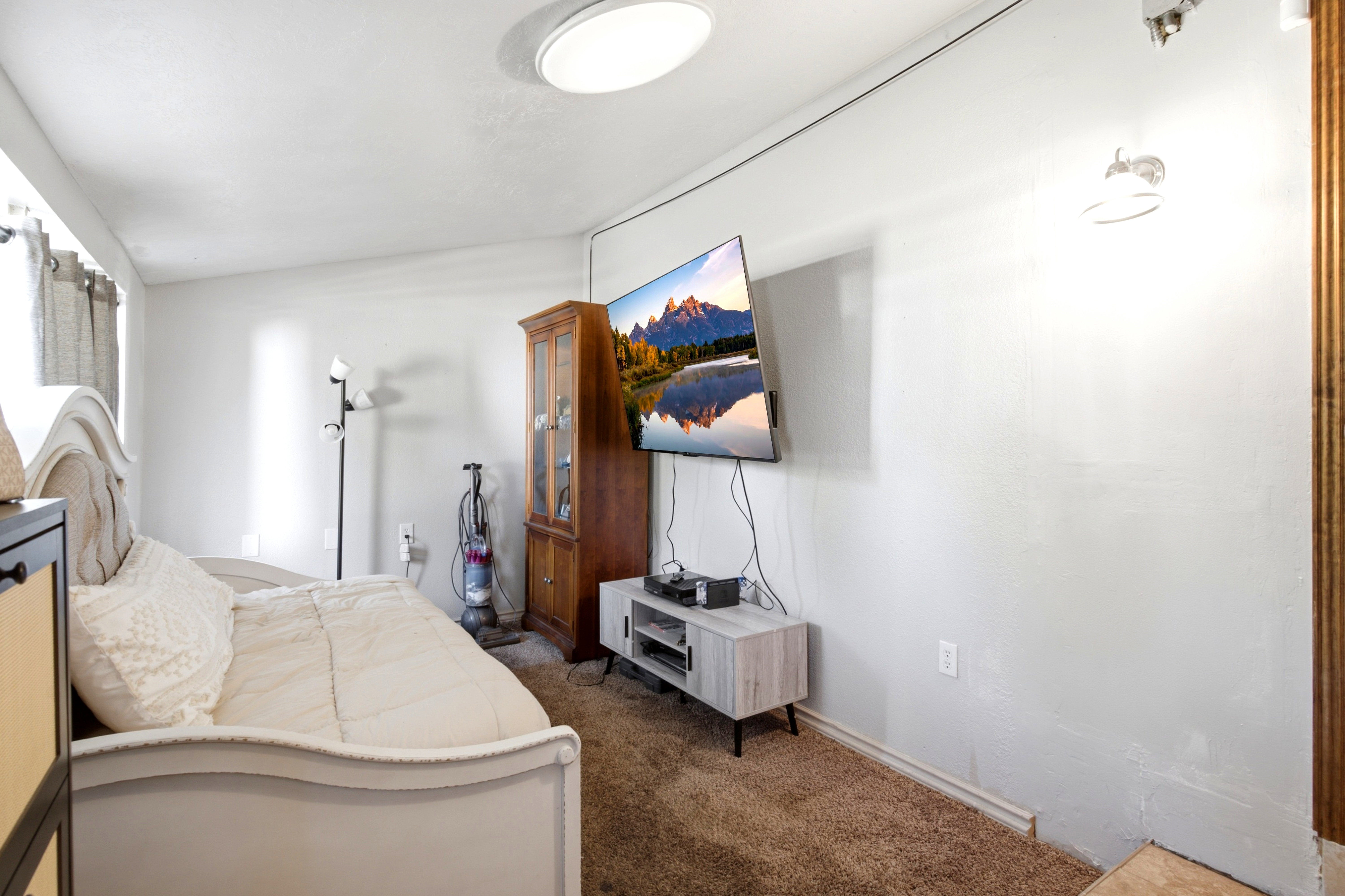 Carpeted room with white walls, featuring a wall-mounted flat-screen television and a ceiling-mounted light fixture