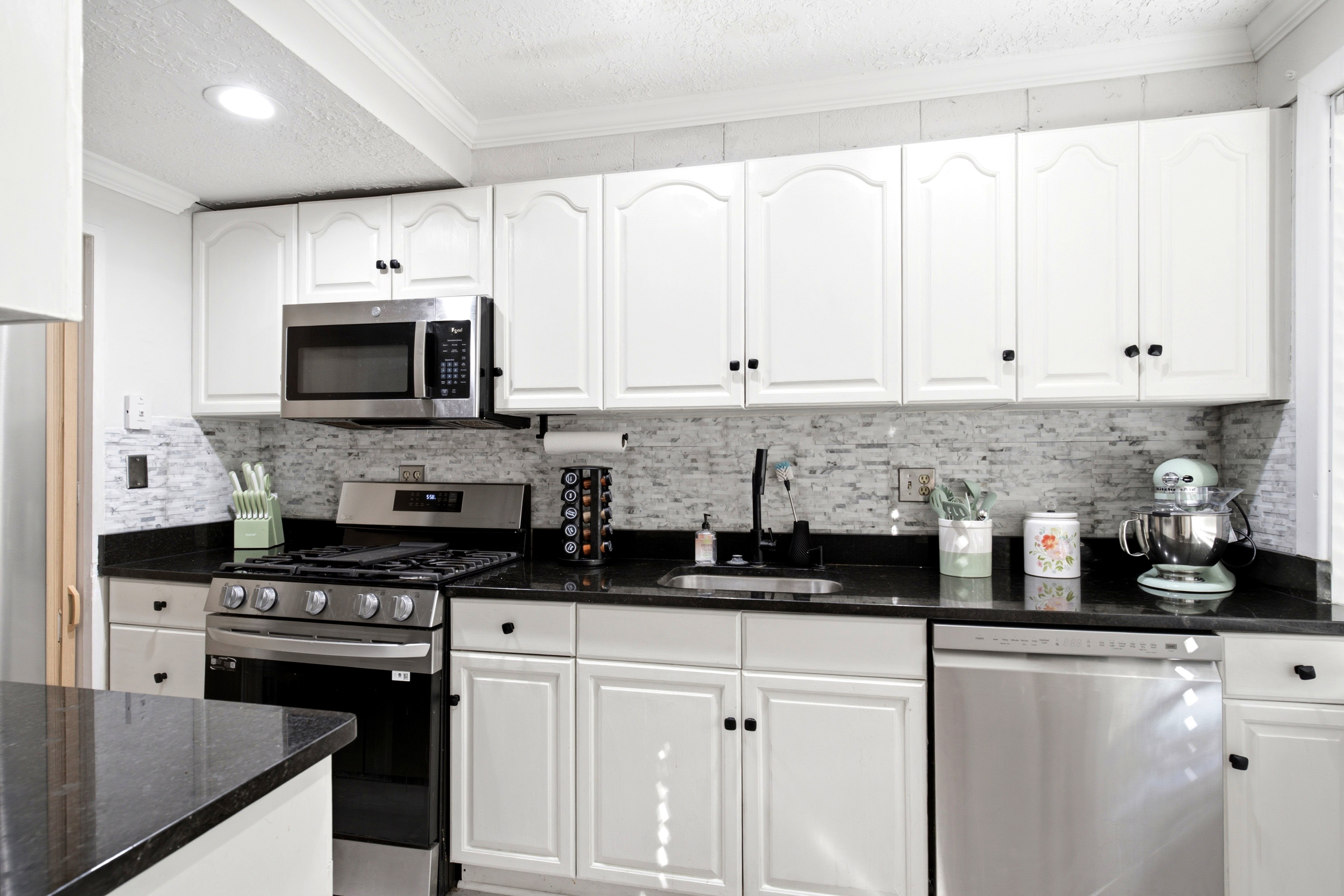 Kitchen featuring white shaker-style cabinetry, black countertops, a mosaic tile backsplash, stainless steel appliances, and a recessed lighting fixture