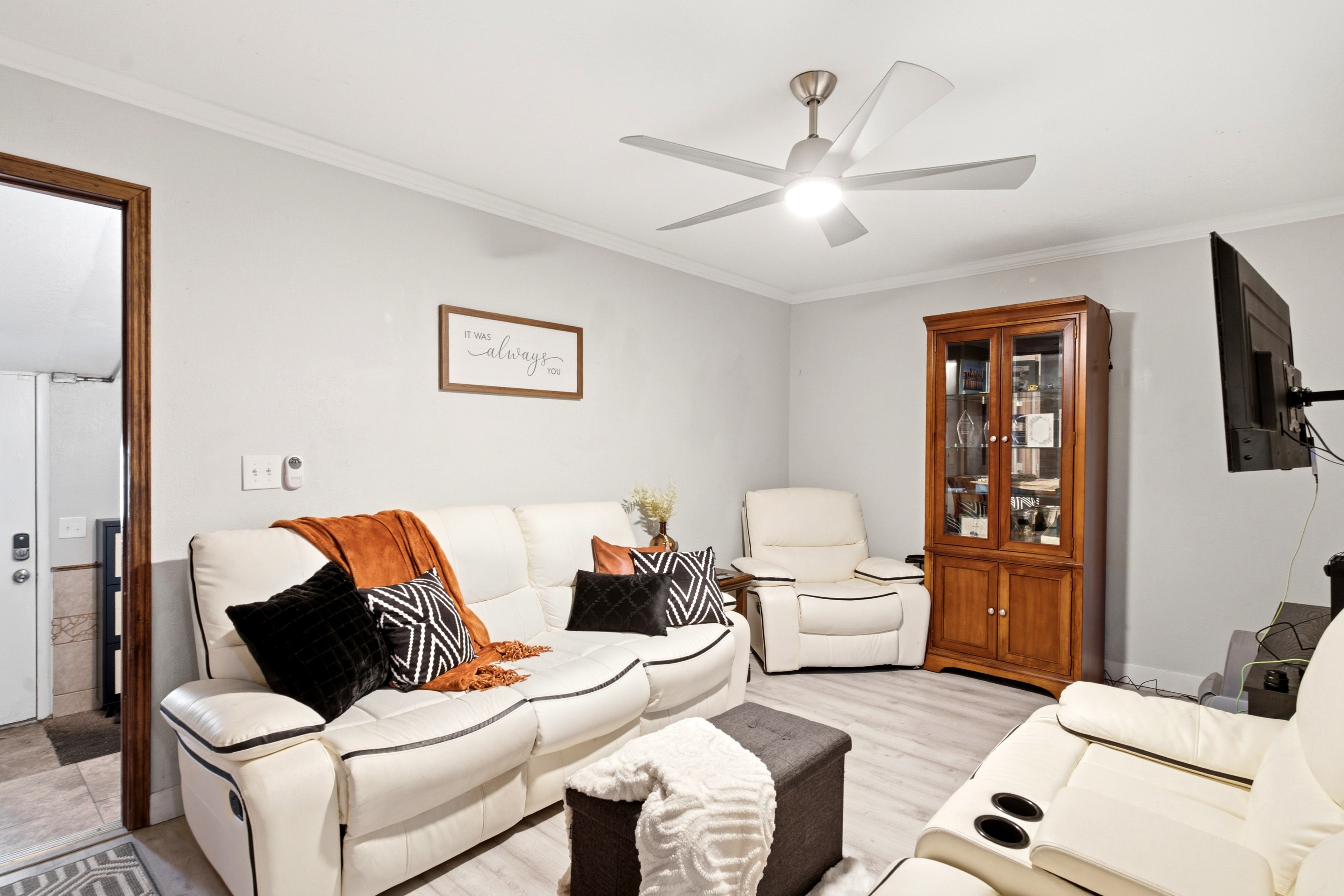 Living area featuring light gray wall paint, white crown molding, wood-finish flooring, a ceiling fan with integrated lighting, and a built-in wood display cabinet
