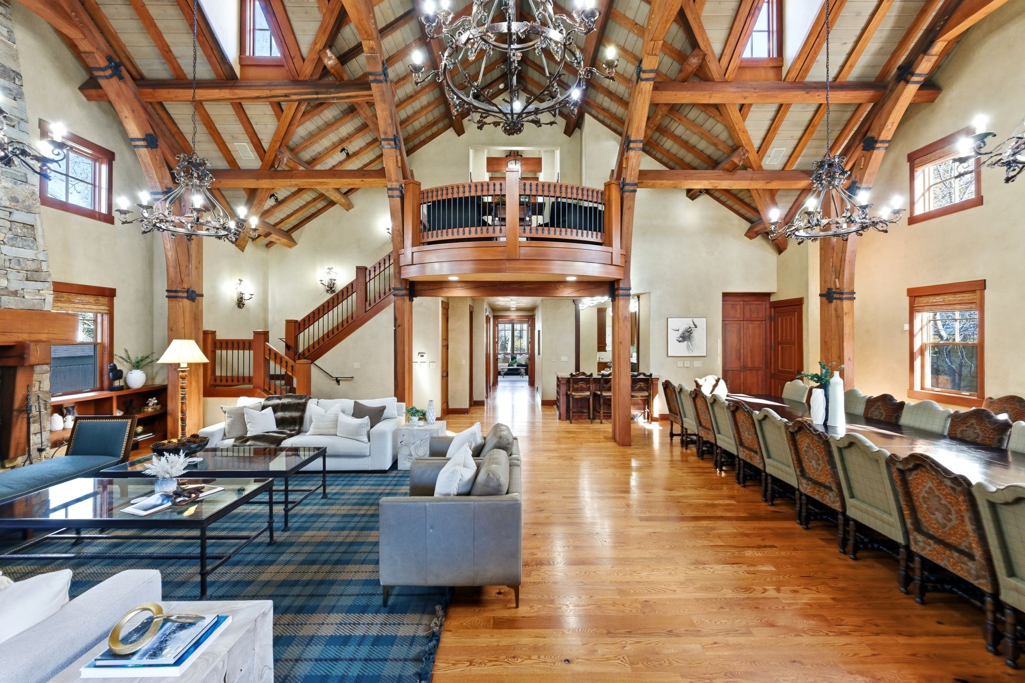 Grand open-concept living space featuring vaulted ceilings, exposed timber beams, a stone fireplace, hardwood flooring, and an upper-level balcony