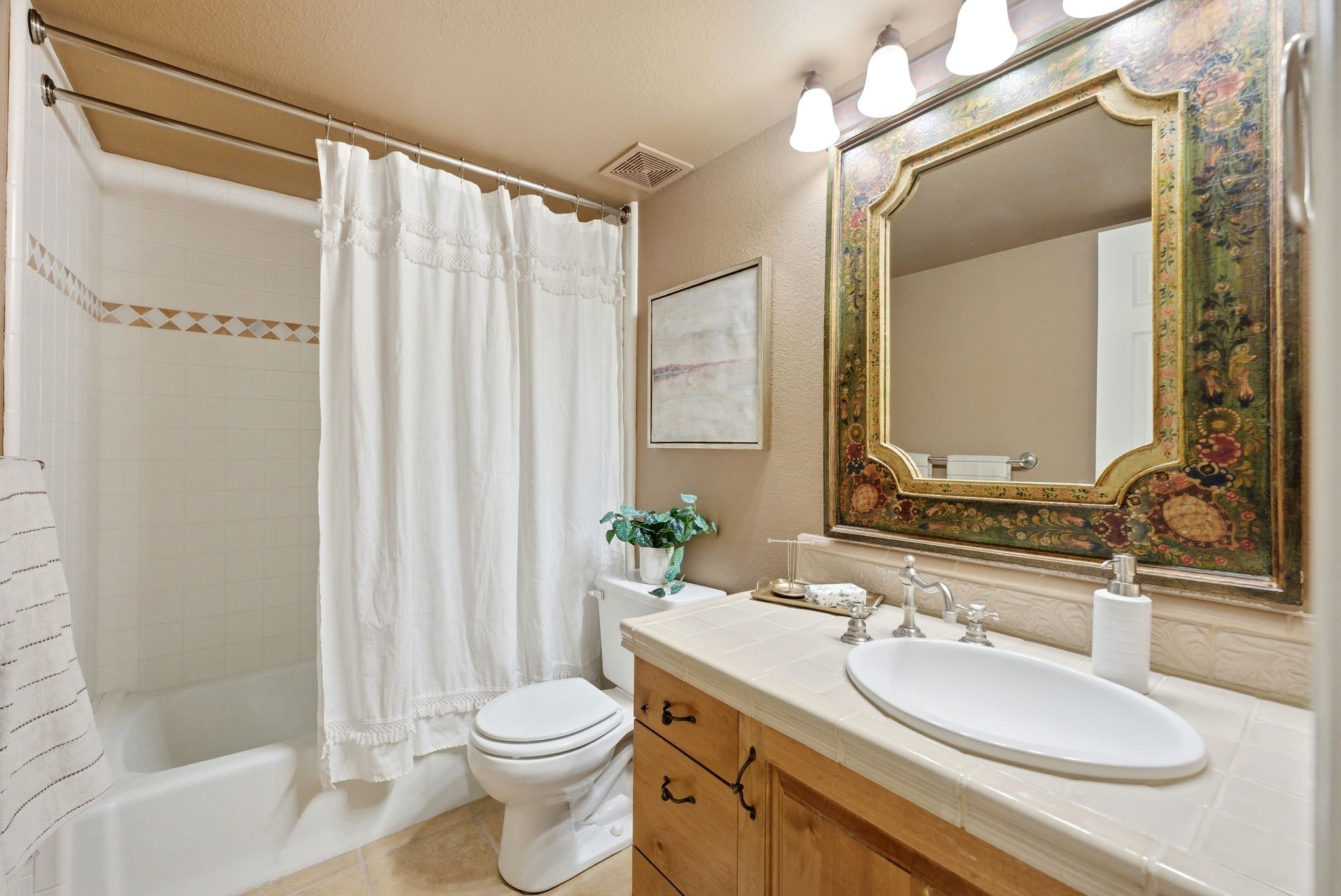 Bathroom featuring a bathtub with a shower, a decorative tile accent band, a toilet, a light wood-finish vanity with panel doors and drawers, and a ceramic tile countertop with a white oval vessel sink