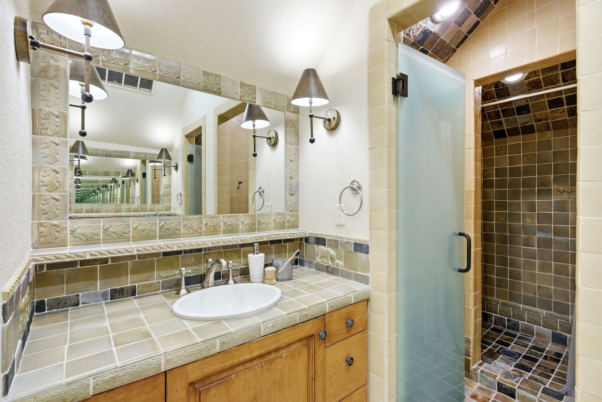 Bathroom featuring a tile vanity countertop with an undermount sink, a large wall-mounted mirror, and a walk-in shower with a frosted glass door
