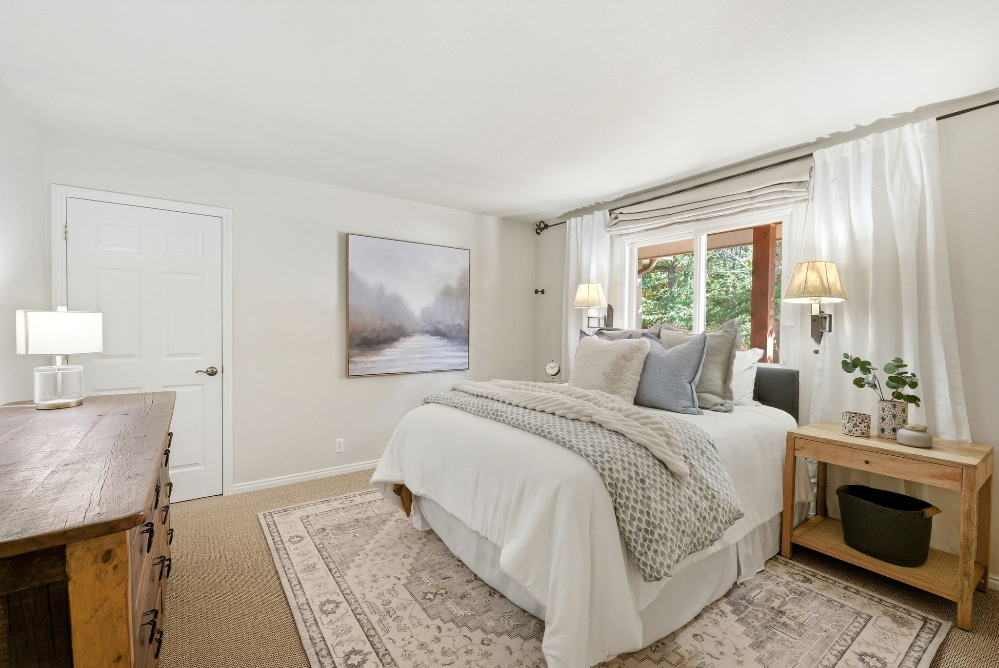 Carpeted bedroom featuring a large window with Roman shades, two wall-mounted sconces, and a paneled door