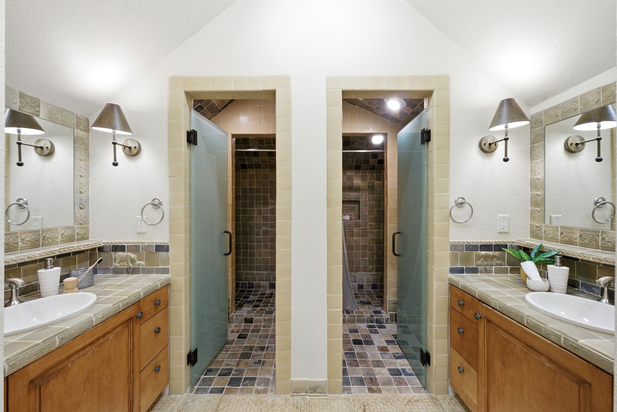 Bathroom featuring dual vanities with tile countertops and integrated sinks, individual showers with glass doors, and tiled flooring