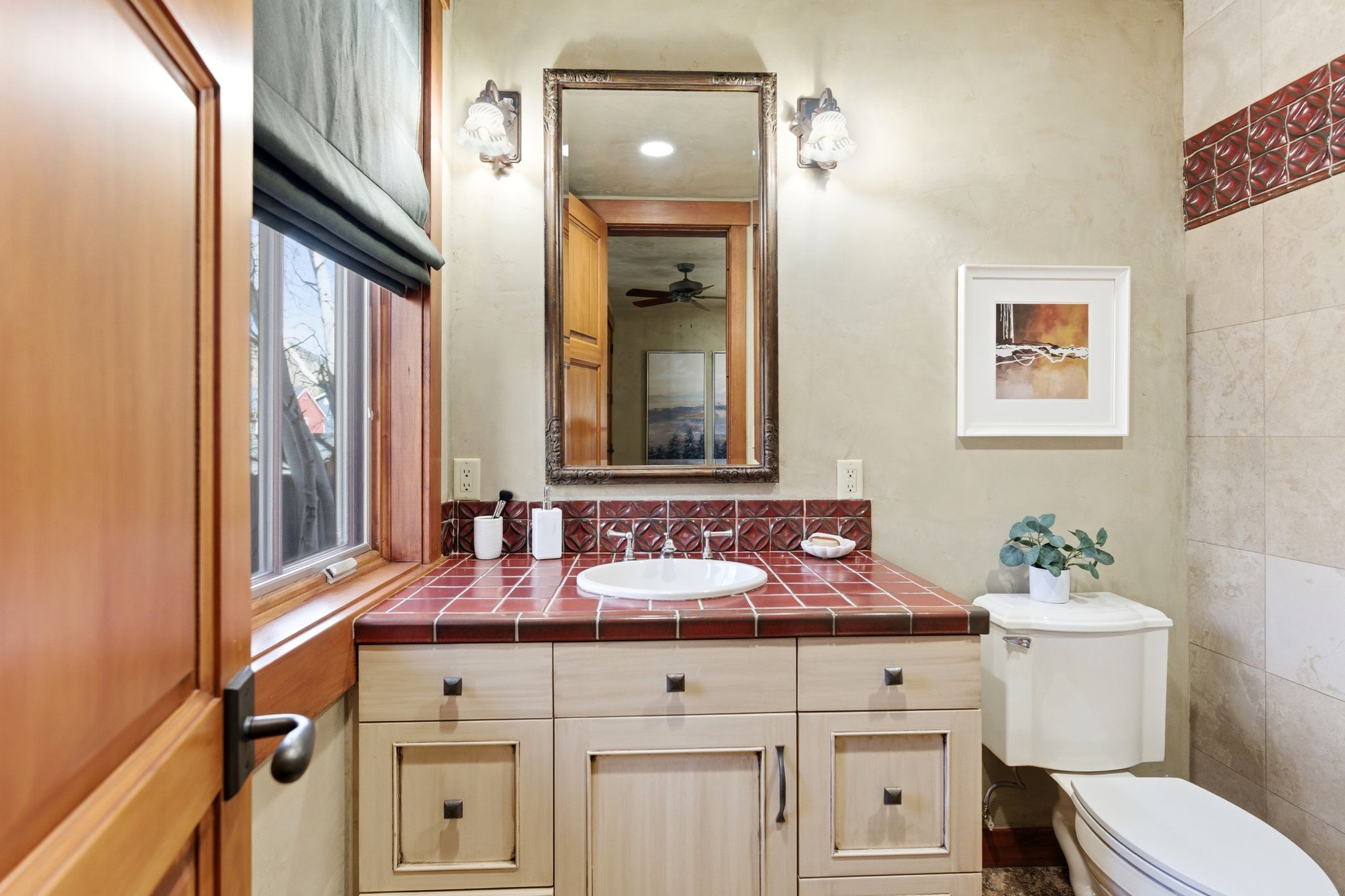 Bathroom featuring a light-toned wood vanity with dark hardware, a red-tiled countertop with an integrated sink, and an ornate framed mirror