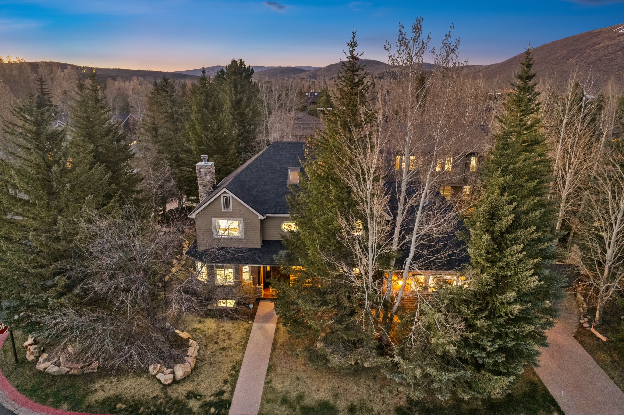 Stucco and stone facade home featuring a prominent stone chimney, dark shingle roof, multiple windows, and a paved walkway leading to the entrance