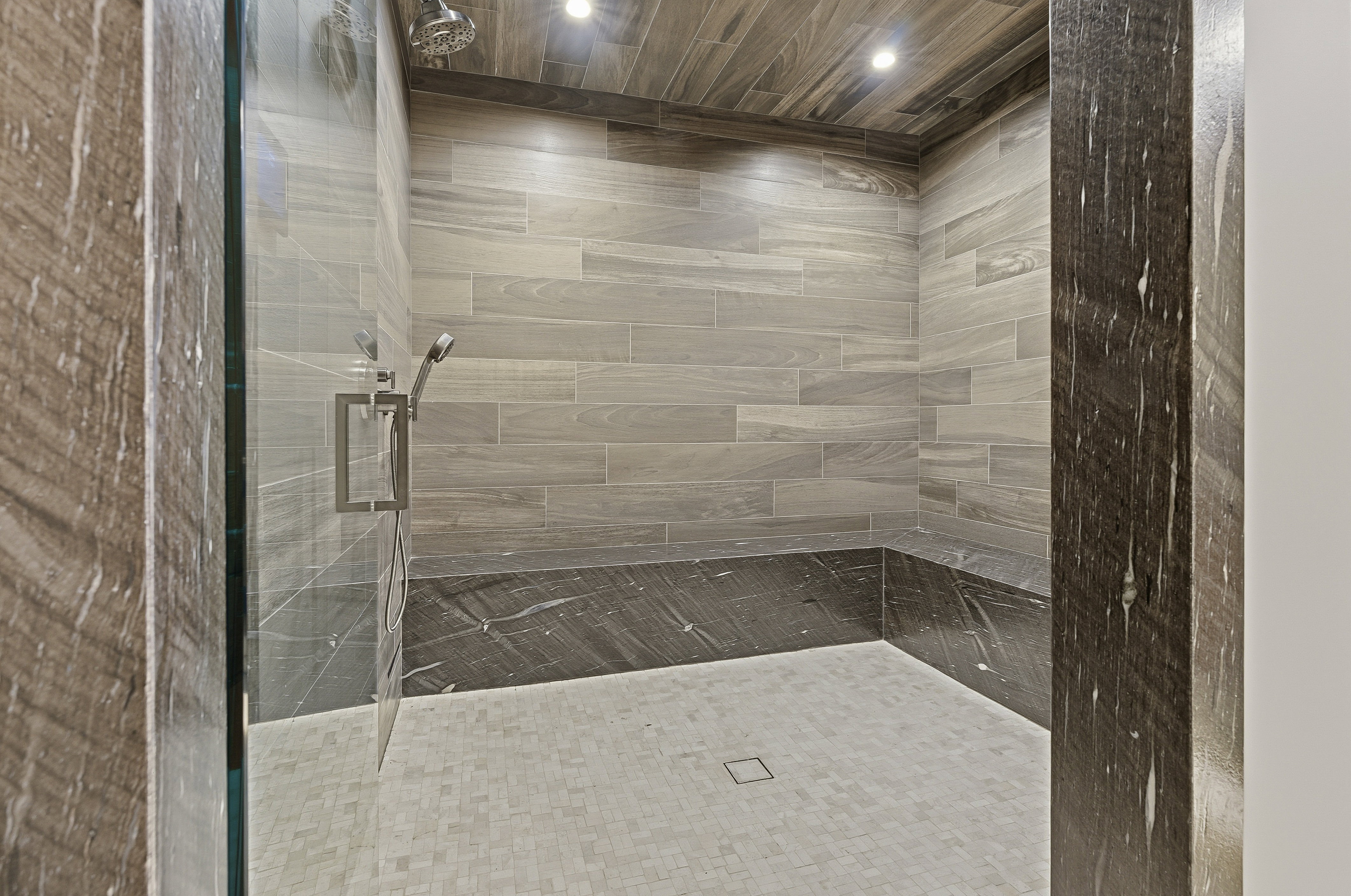 Walk-in shower featuring a glass enclosure, a built-in bench with dark stone, light wood-finish ceiling, and light tile walls