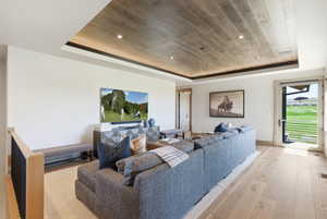 Living space featuring a wood-finish tray ceiling with recessed lighting, light wood-finish flooring, a wall-mounted flat-screen television, and a glass-paneled door opening to a green expanse