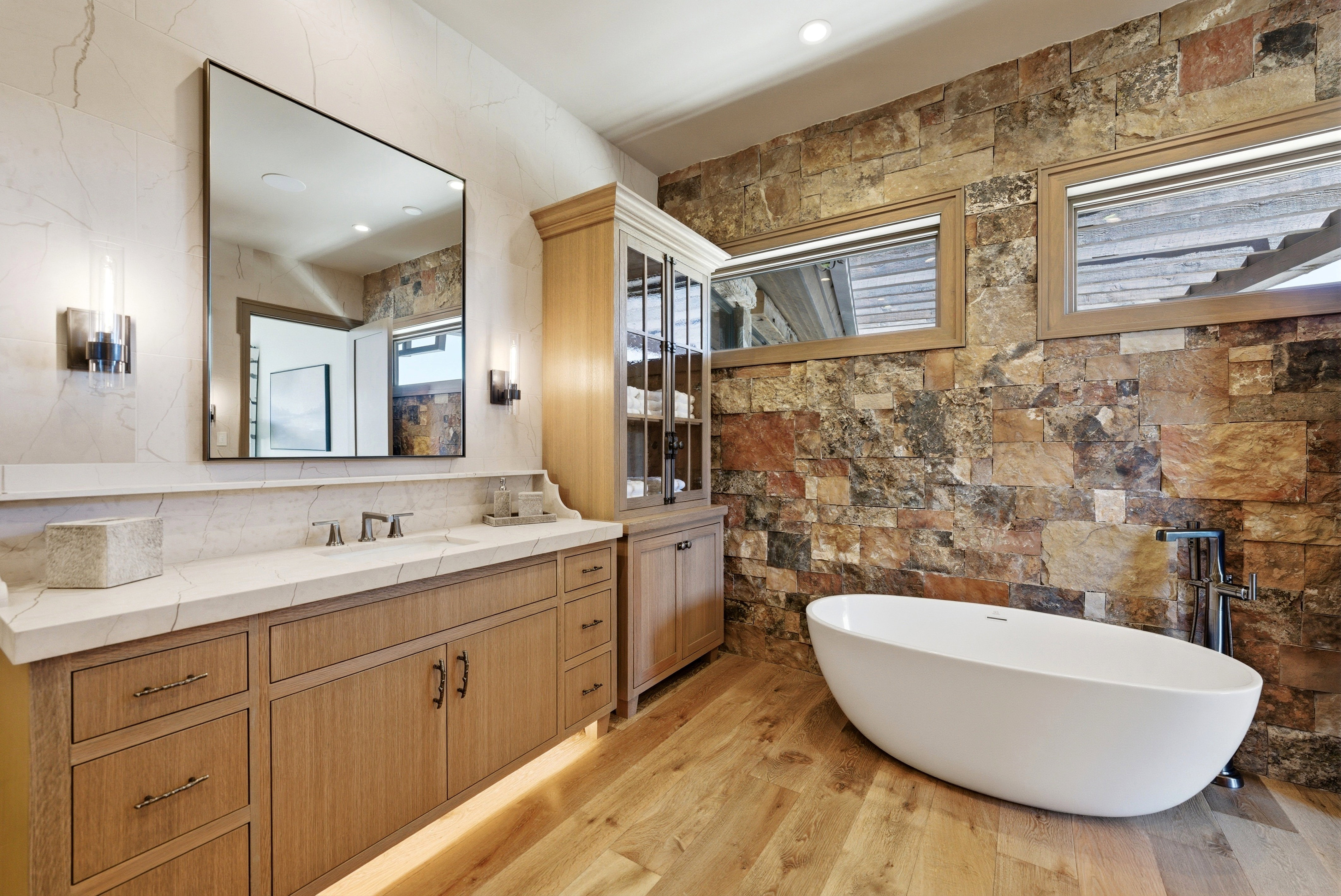Freestanding soaking tub with a stone accent wall, wood-finish flooring, a double vanity with a light-toned countertop, and integrated under-cabinet lighting