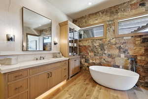 Freestanding soaking tub with a stone accent wall, wood-finish flooring, a double vanity with a light-toned countertop, and integrated under-cabinet lighting