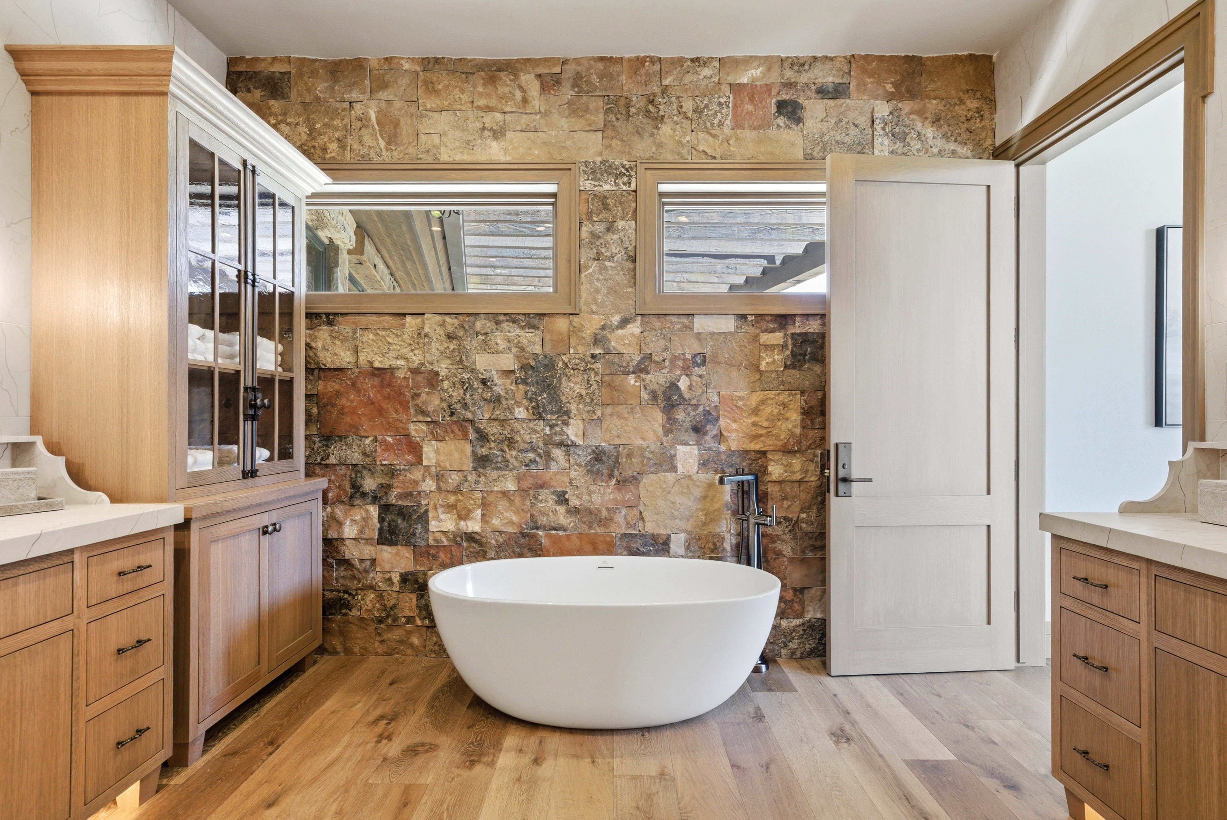Freestanding soaking tub set against a stone accent wall, complemented by light wood-finish flooring
