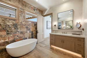 Bathroom featuring a freestanding soaking tub, stone accent wall, wood-finish flooring, a wood vanity with a white countertop, and a large wall-mounted mirror
