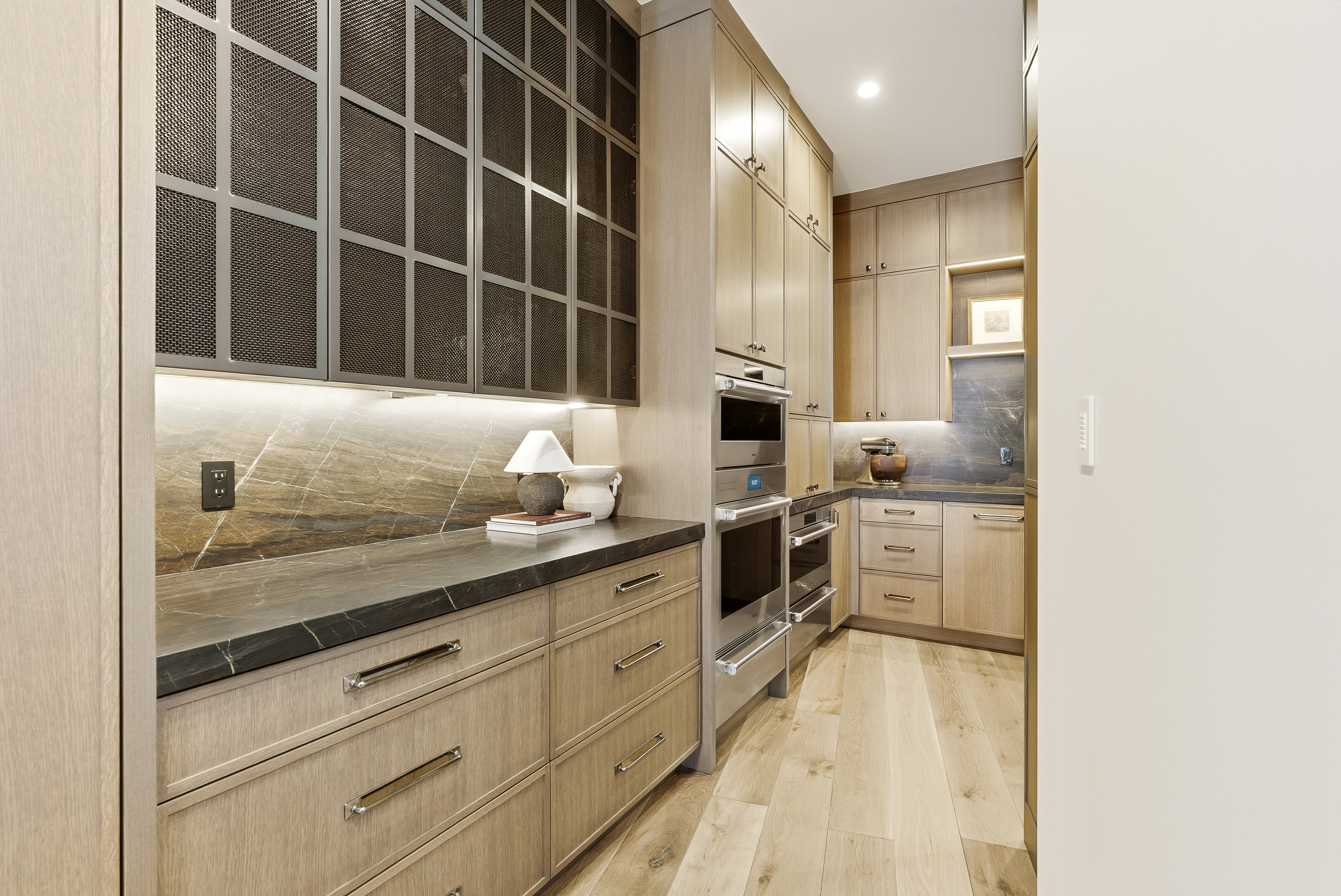 Custom cabinetry with metal mesh inserts, under-cabinet lighting, a stone slab backsplash, dark stone countertops, and stainless steel built-in appliances