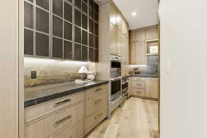 Custom cabinetry with metal mesh inserts, under-cabinet lighting, a stone slab backsplash, dark stone countertops, and stainless steel built-in appliances