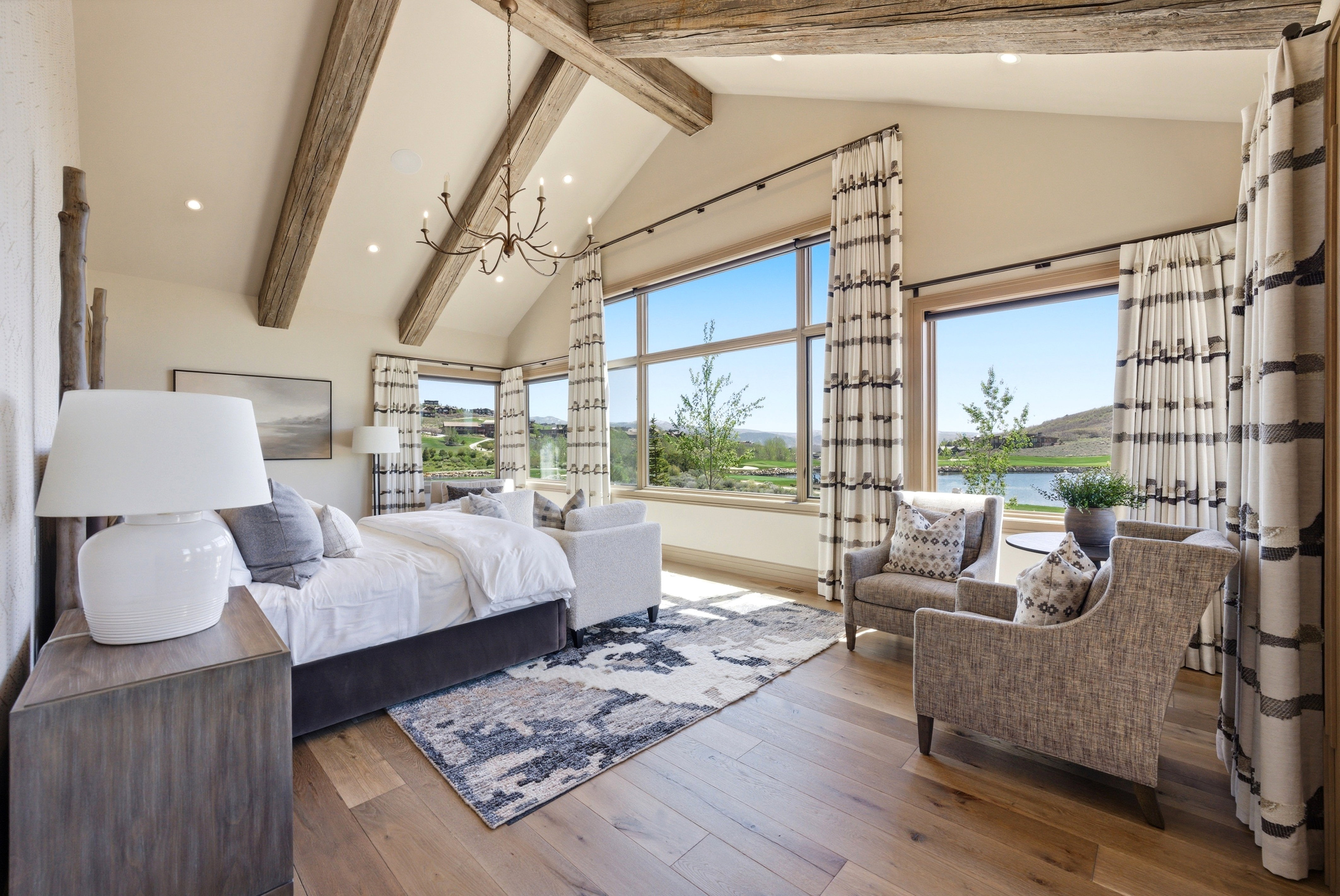 Vaulted ceilings with exposed wood beams define this spacious room, featuring large windows with verdant landscaping and water visible
