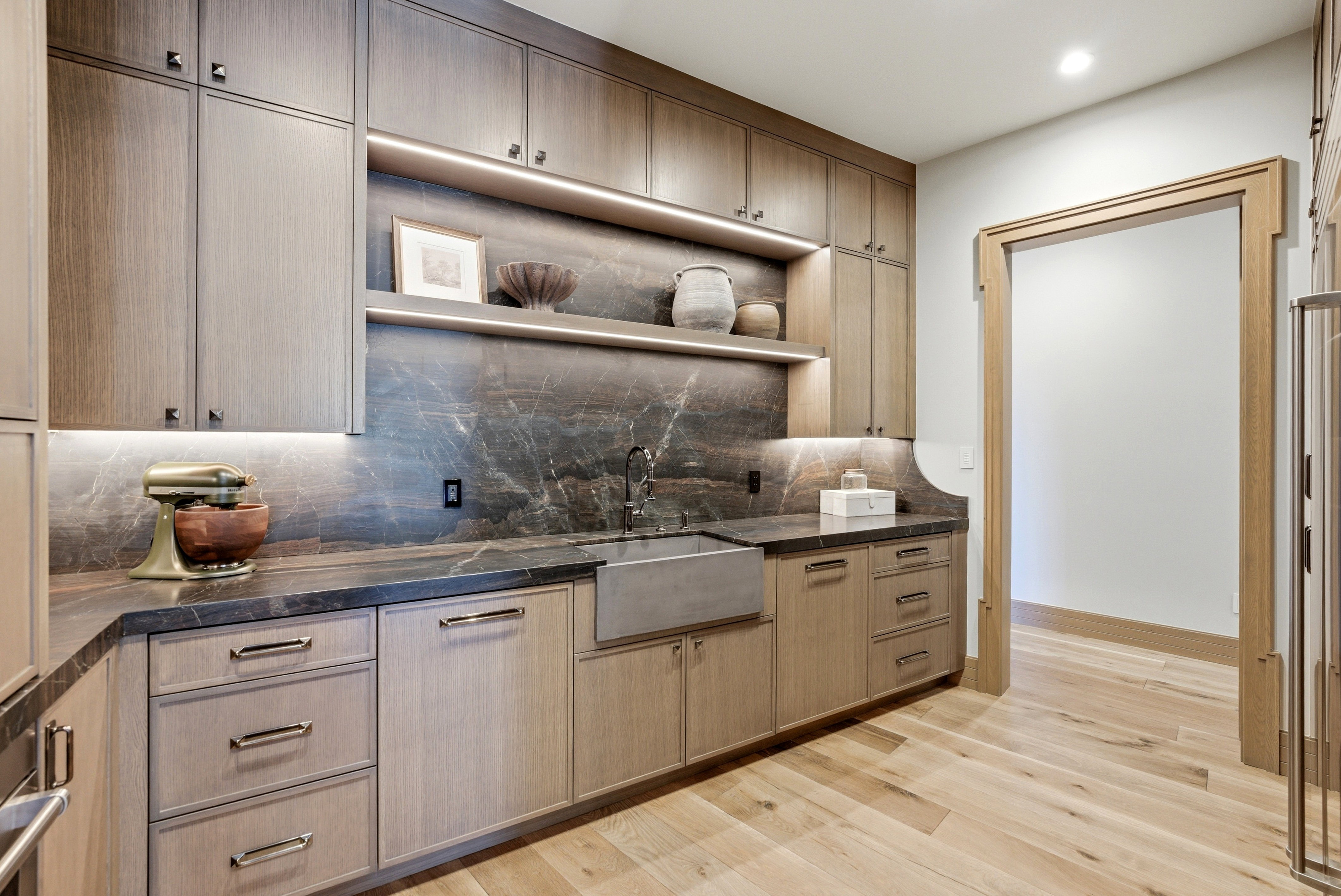 Custom cabinetry with integrated under-cabinet lighting, a deep farmhouse-style sink, and a dark stone countertop with a matching backsplash