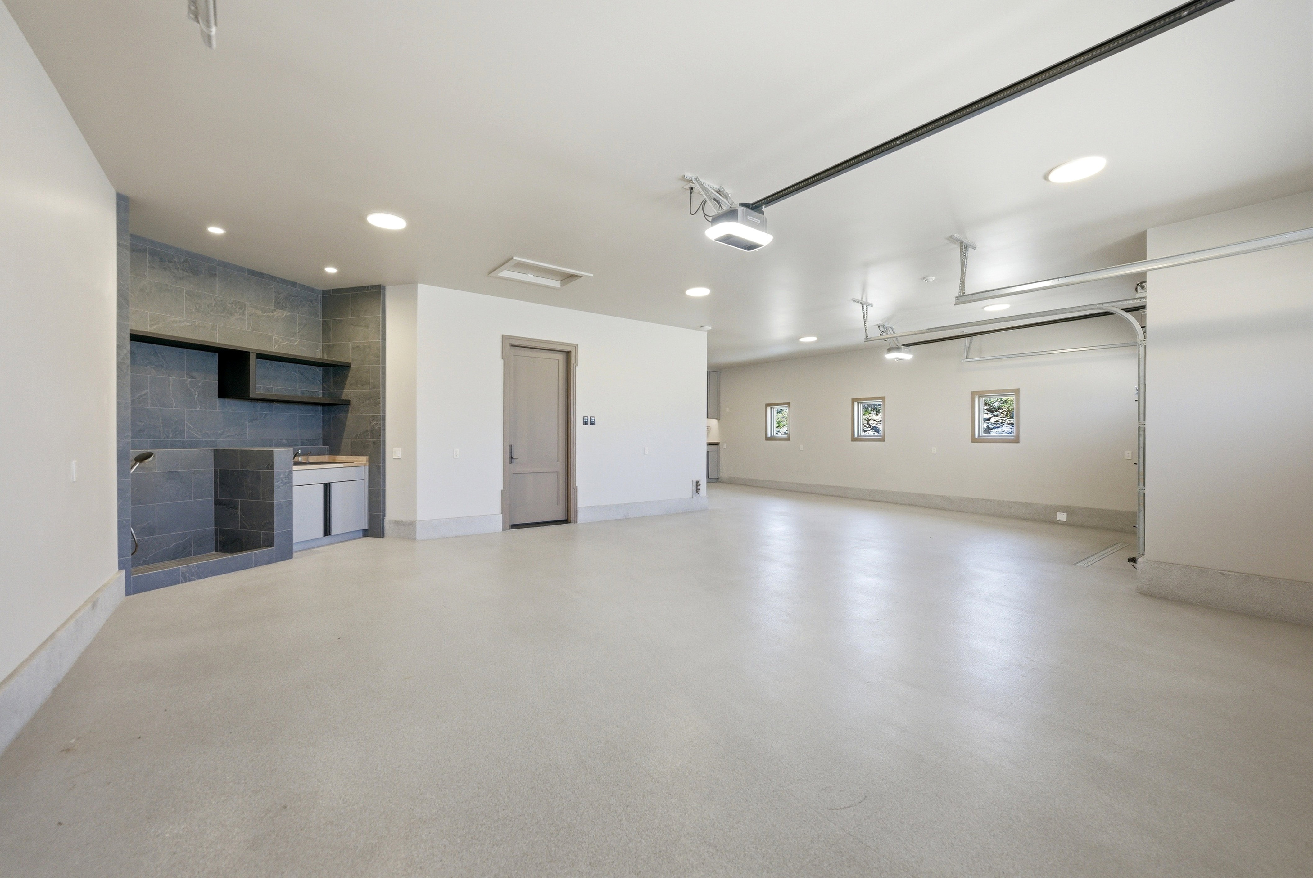 Spacious garage featuring a tiled utility sink area with built-in shelving, light-colored concrete flooring, recessed ceiling lighting, and an automatic garage door opener