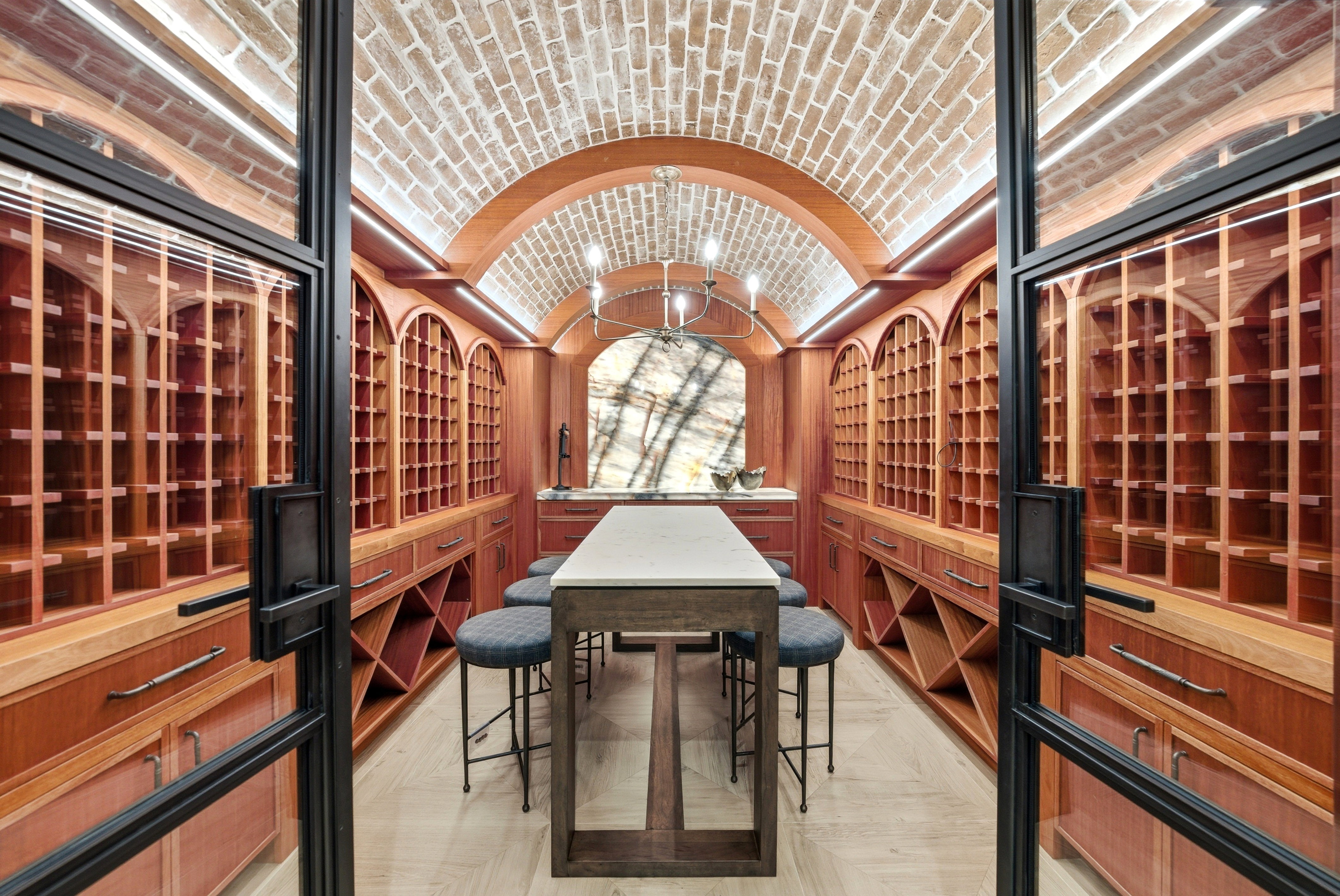 Custom wine cellar featuring an arched brick ceiling, extensive wood-finish cabinetry, built-in wine racks, an illuminated stone accent wall, and a central tasting table