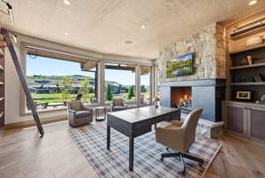Expansive windows showcasing hillside landscaping, a stone-clad fireplace with a dark metal mantel, light wood-finish flooring, built-in shelving, and a textured ceiling finish