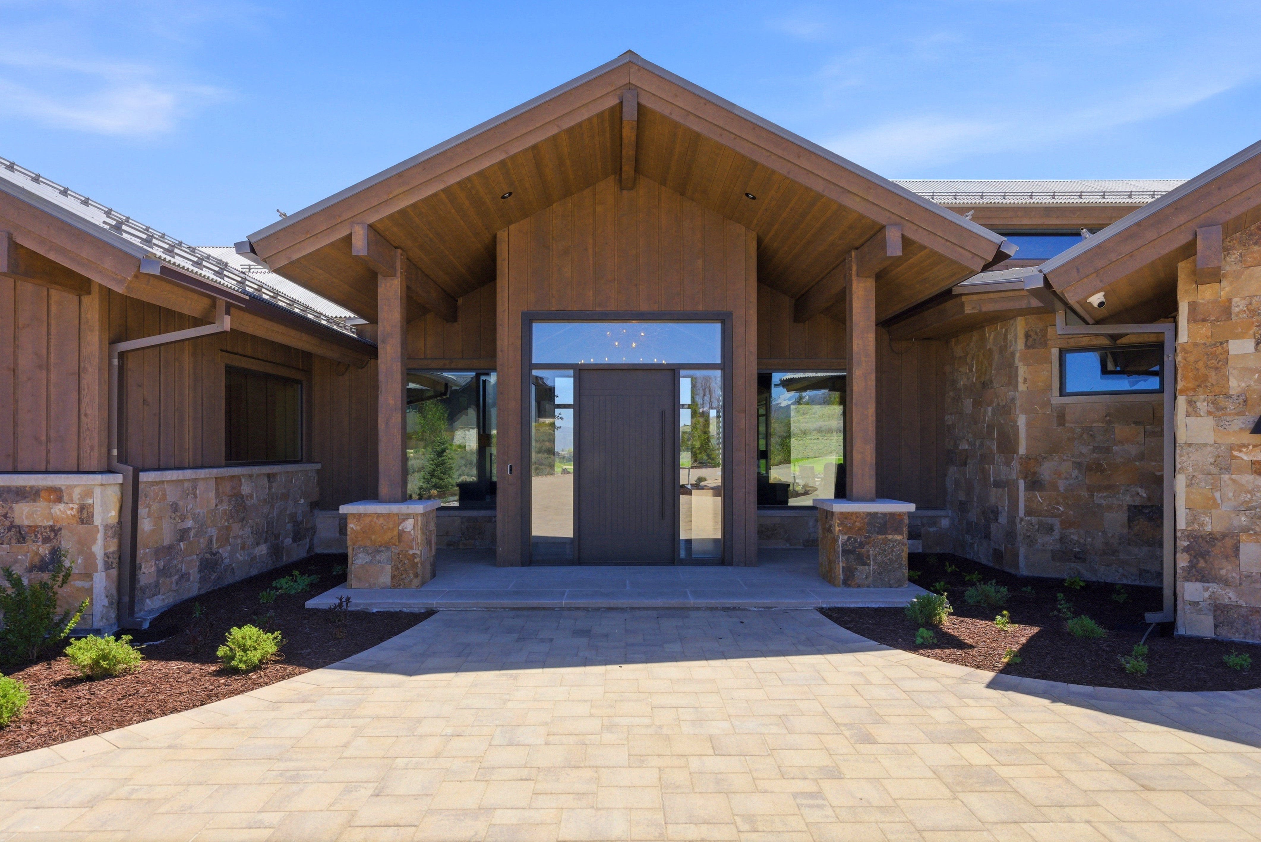 Grand entrance featuring a double door with sidelights, timber frame porch, natural stone veneer, vertical wood siding, and a paver driveway
