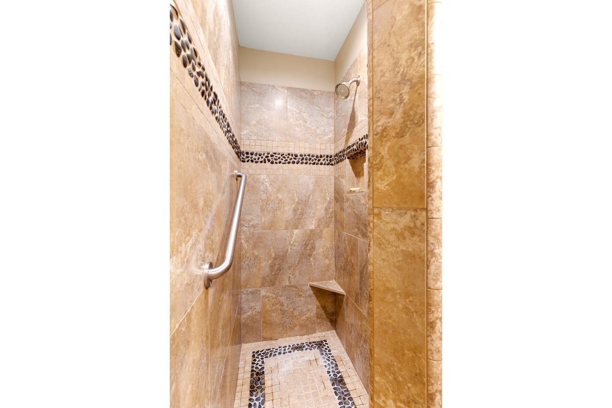 Walk-in shower featuring natural stone-look wall tiles, pebble-detail accent bands, a built-in shower bench, and a brushed nickel grab bar