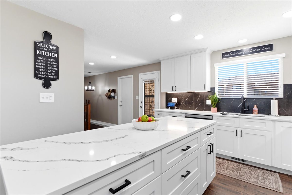 Modern kitchen featuring a large center island with a white veined countertop, white cabinetry with matte black hardware, dark stone-patterned backsplash, and recessed lighting