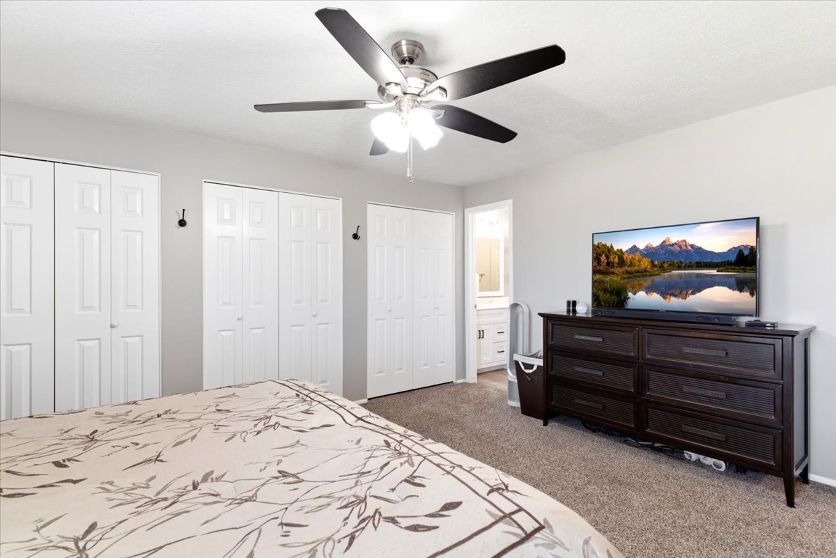Carpeted room featuring multiple bifold closet doors, a ceiling fan with integrated lighting, and direct access to a bathroom with a vanity and mirror
