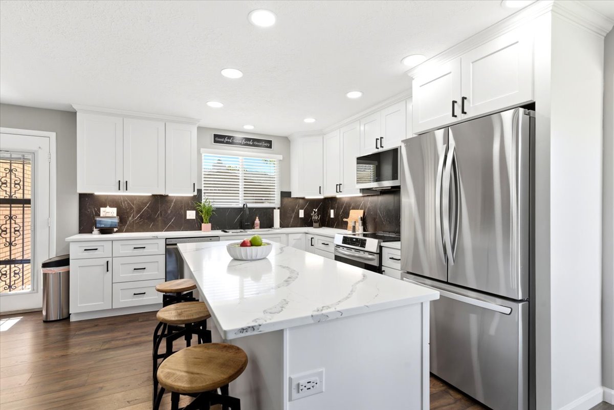 Modern kitchen featuring a large central island with a waterfall-edge countertop, white shaker-style cabinetry, stainless steel appliances, and wood-finish flooring