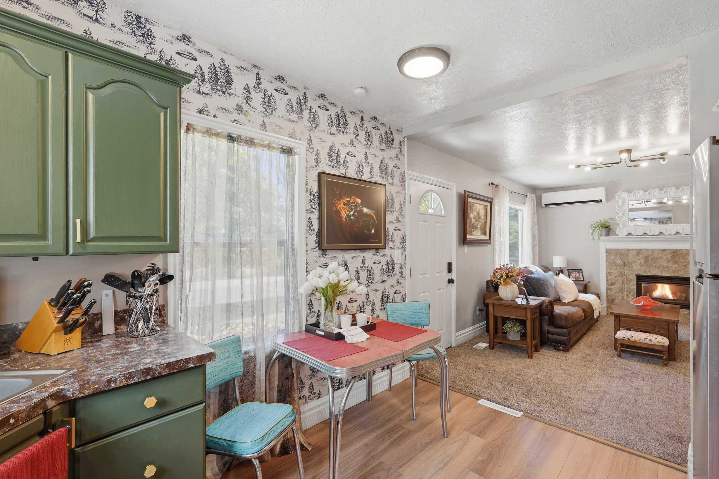 Open-concept living space featuring wood-finish flooring, a stone-faced fireplace, a ductless mini-split system, green cabinetry, and patterned wallpaper