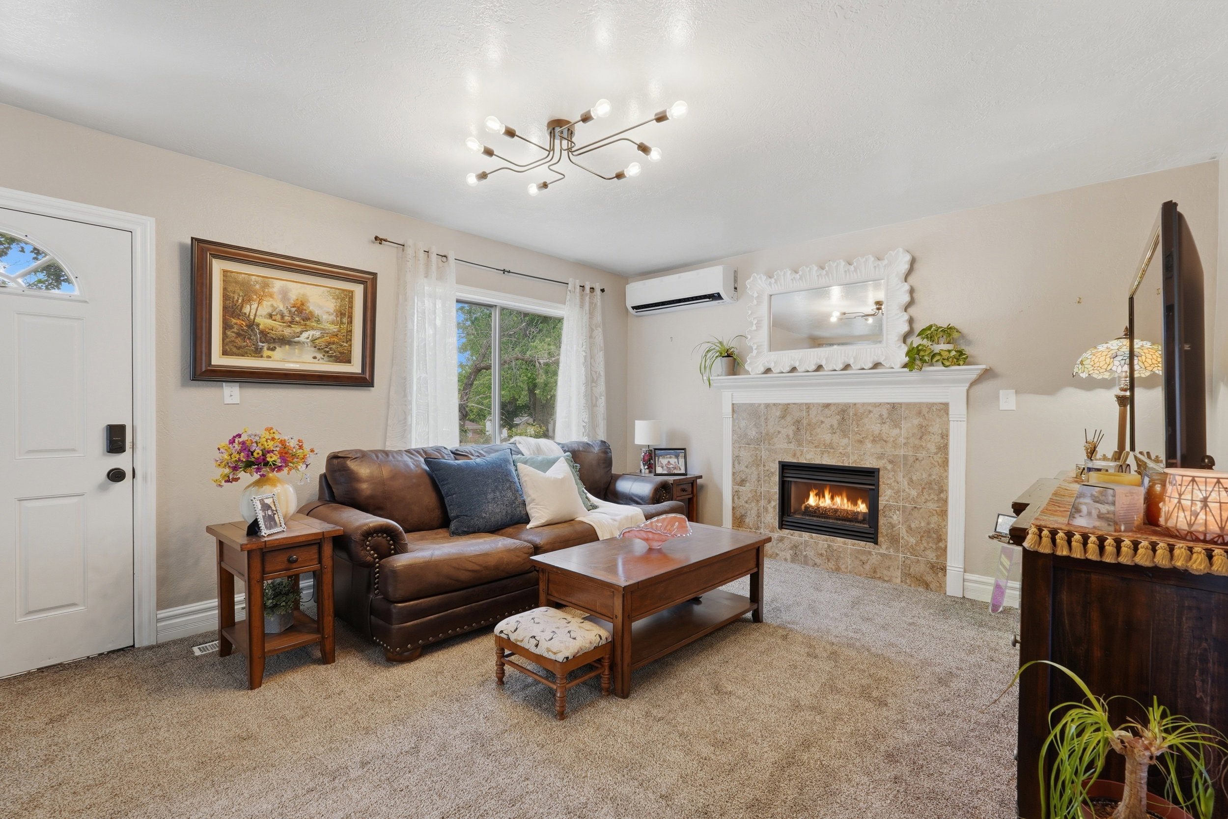 Living room featuring a gas fireplace with a tiled surround and white mantel, a wall-mounted mini-split unit, a contemporary ceiling light fixture, a window with exterior views, and neutral-toned carpeting