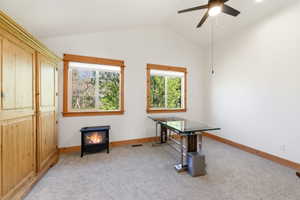 Vaulted ceilings with a ceiling fan, two double-hung windows with natural wood trim, wall-to-wall carpeting, and a freestanding fireplace