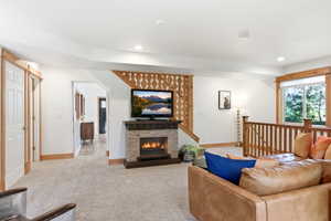 Living area featuring a stone-clad fireplace, recessed lighting, carpet flooring, a large window, and wooden staircase railing