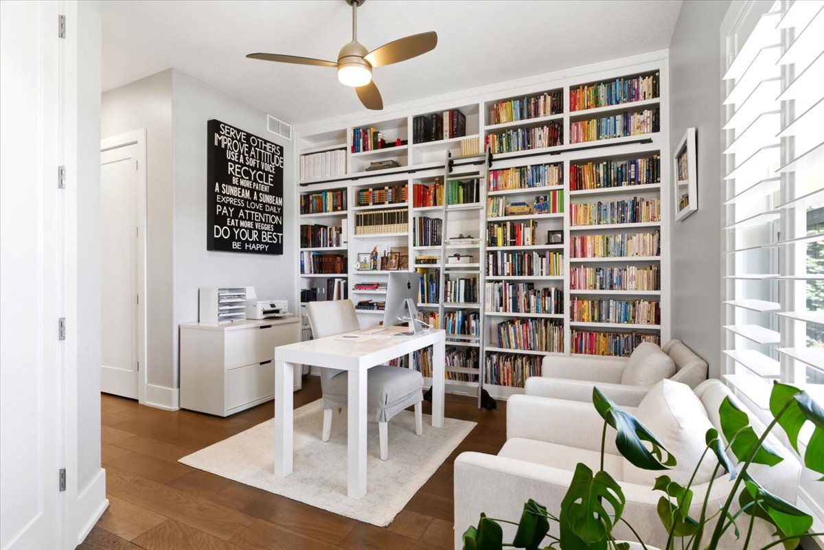 Dedicated office space featuring extensive built-in shelving, a rolling library ladder, wood-finish flooring, and a ceiling fan