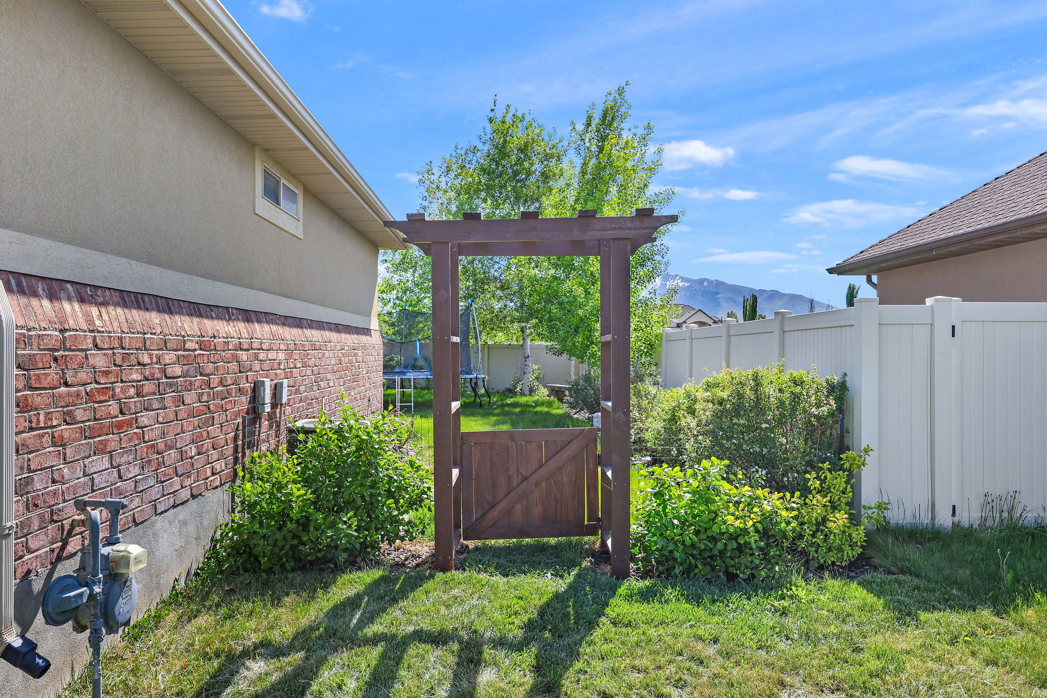Landscaped backyard featuring a wooden arbor with a privacy gate, manicured lawn, established shrubs, and a neutral-toned vinyl fence