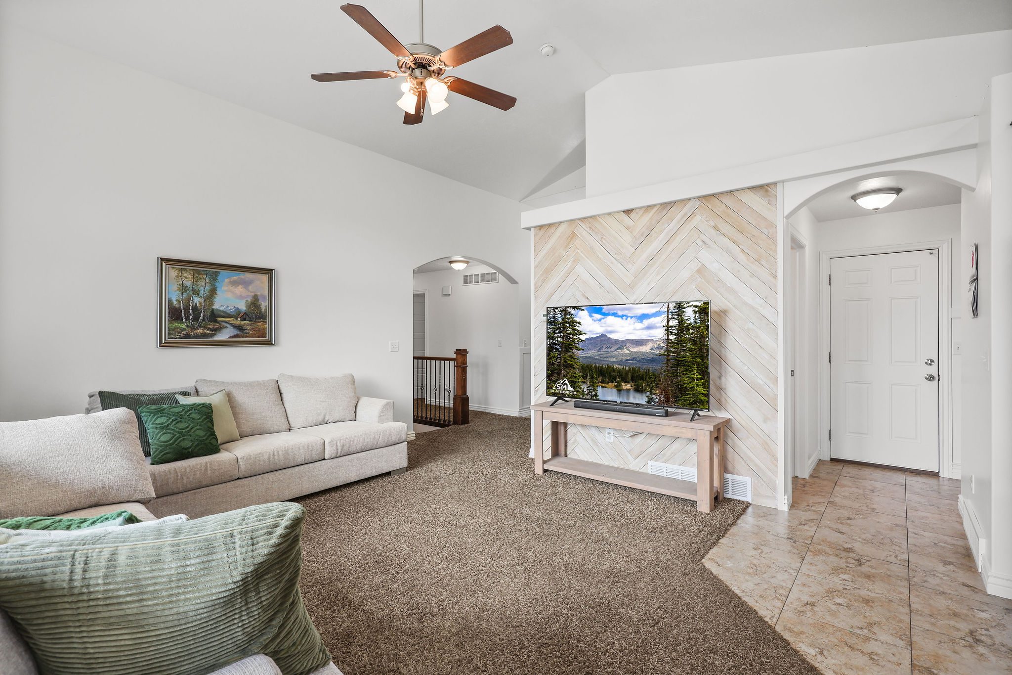 Vaulted ceiling living area featuring a ceiling fan with integrated lighting, a decorative chevron wood-finish accent wall, and a tiled entryway