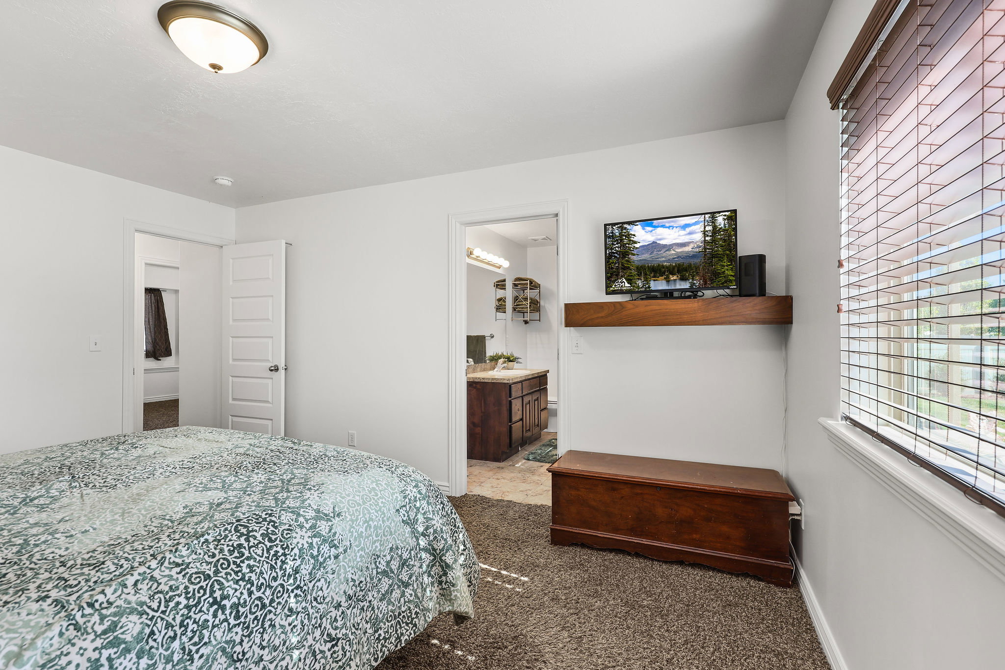 Carpeted bedroom featuring a window with blinds, a wood-finish floating shelf, a ceiling-mounted light fixture, and white trim