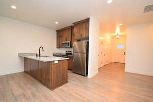 Kitchen featuring an island with a built-in sink, granite countertops, wood-finish cabinetry, stainless steel appliances, and wood-finish flooring