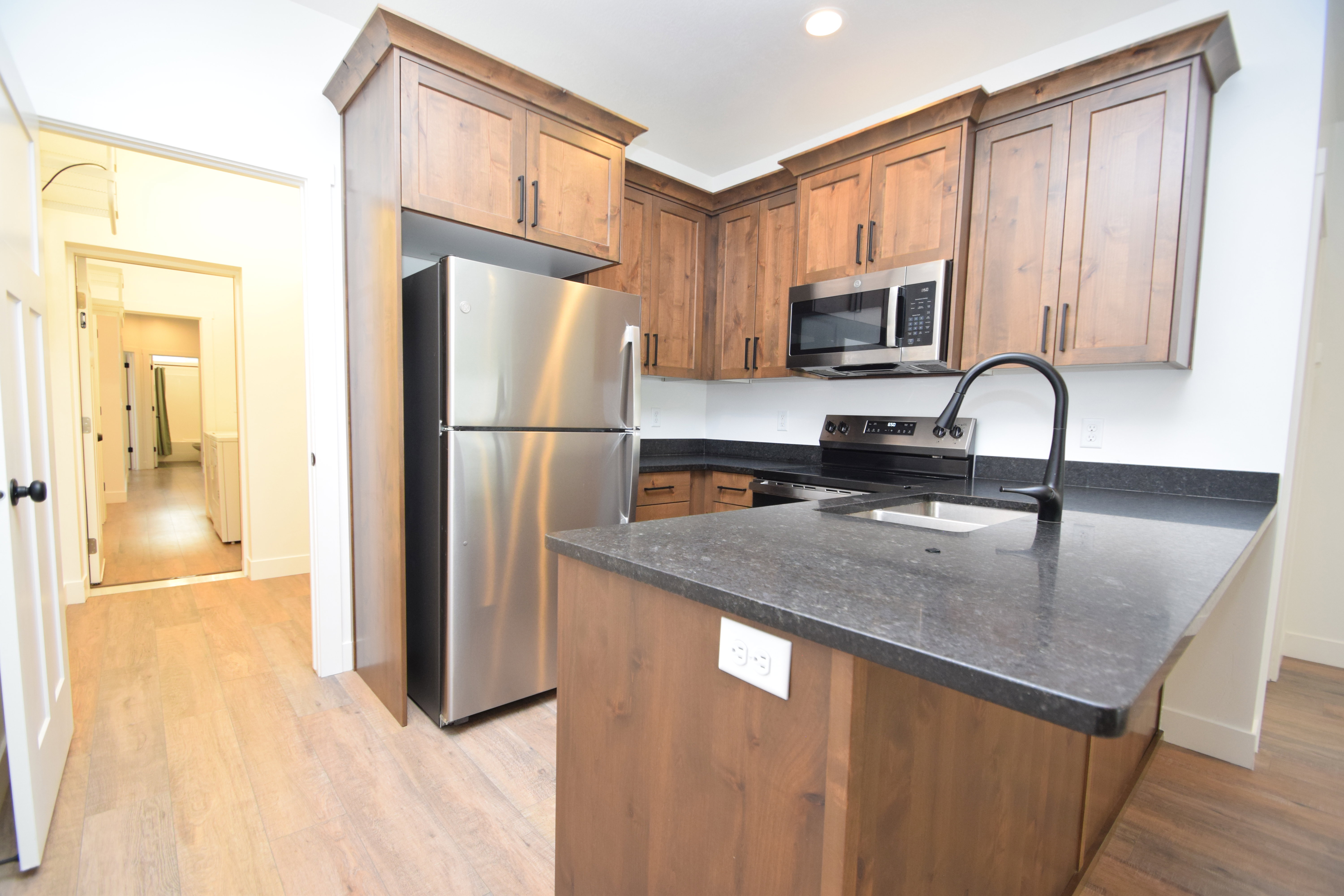 Kitchen featuring wood-finish cabinetry, stainless steel appliances, dark countertops, a kitchen island with an undermount sink, and light wood-finish flooring