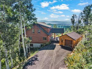 Log-style main residence with covered entry, separate garage structure, paved driveway, and mature trees on a sloped lot