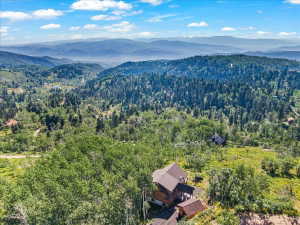Expansive mountain landscape featuring a prominent residential structure with multiple levels and a brown roof, surrounded by dense green forestry