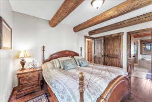 Bedroom featuring exposed wood ceiling beams, wood-finish flooring, wood paneled doors, and an en-suite bathroom with a built-in tub