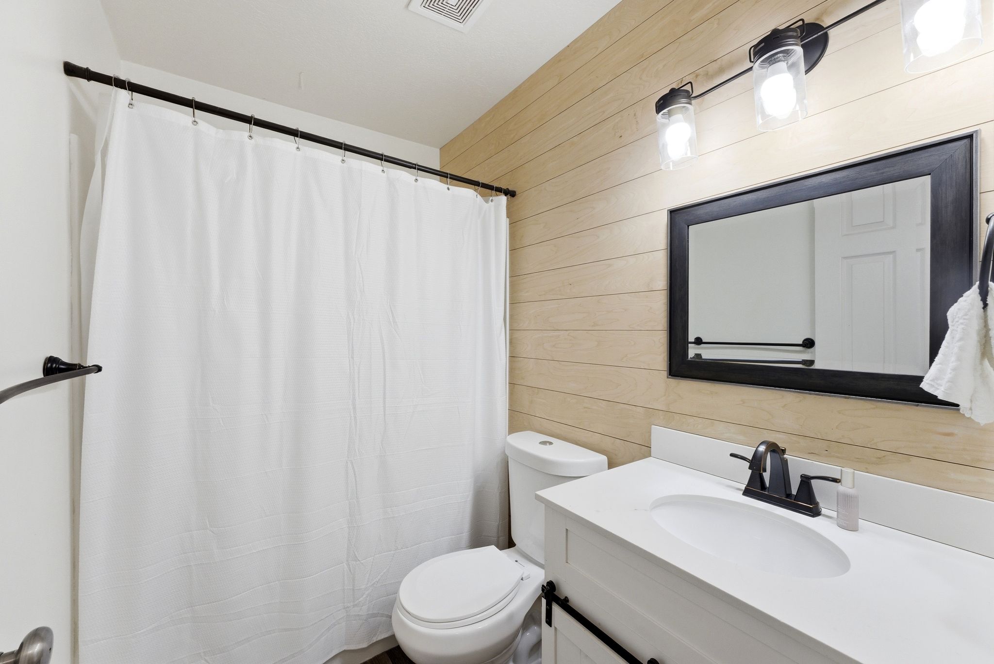 Bathroom with wood-finish accent wall, white vanity with integrated sink, black framed mirror, and black wall sconce lighting