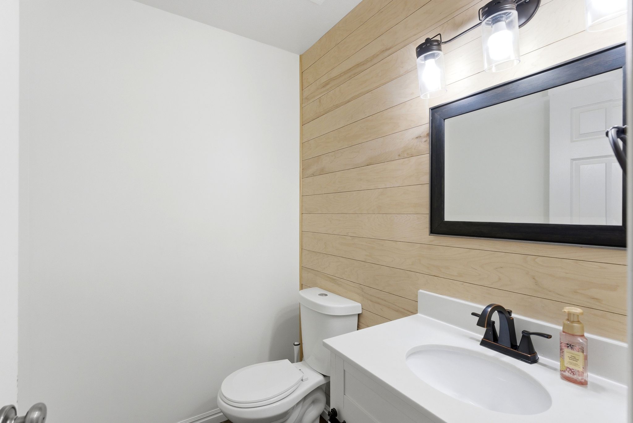 Powder room featuring a wood-finish accent wall, white vanity with integrated sink, oil-rubbed bronze faucet, and a rectangular framed mirror with overhead lighting
