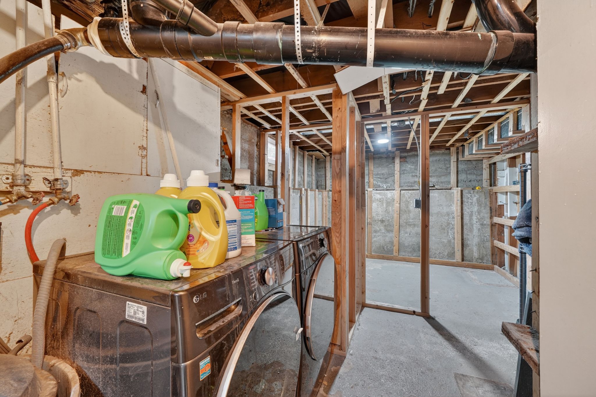 Unfinished basement area featuring exposed wood framing, concrete flooring, exposed overhead piping, and a dedicated laundry appliance space