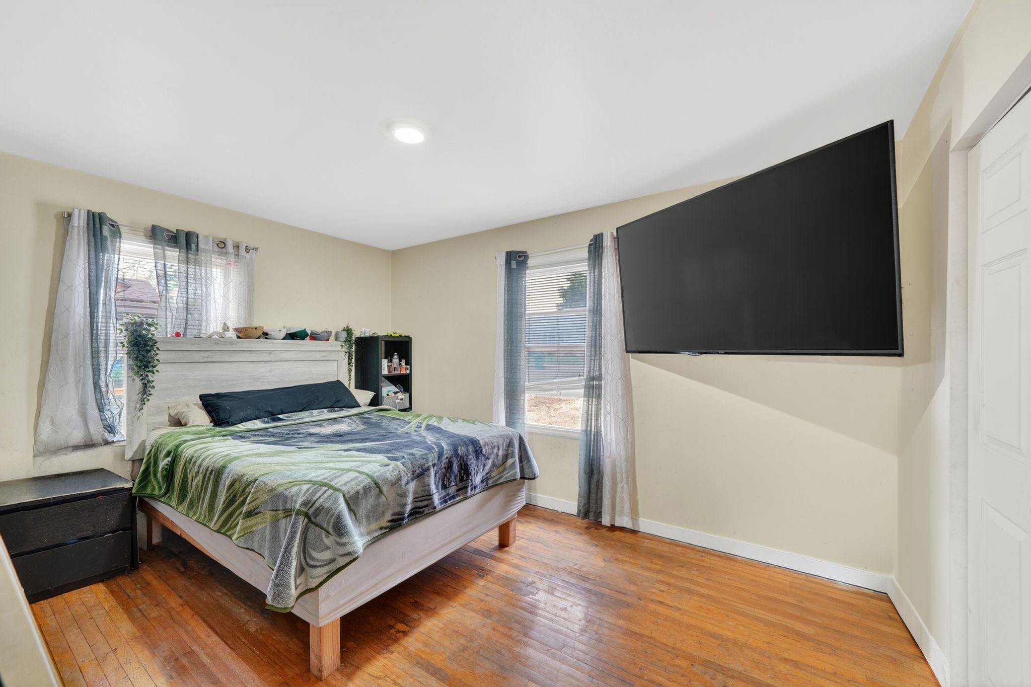 Bedroom featuring hardwood flooring, two windows, a white paneled door, a recessed ceiling light, and a wall-mounted television