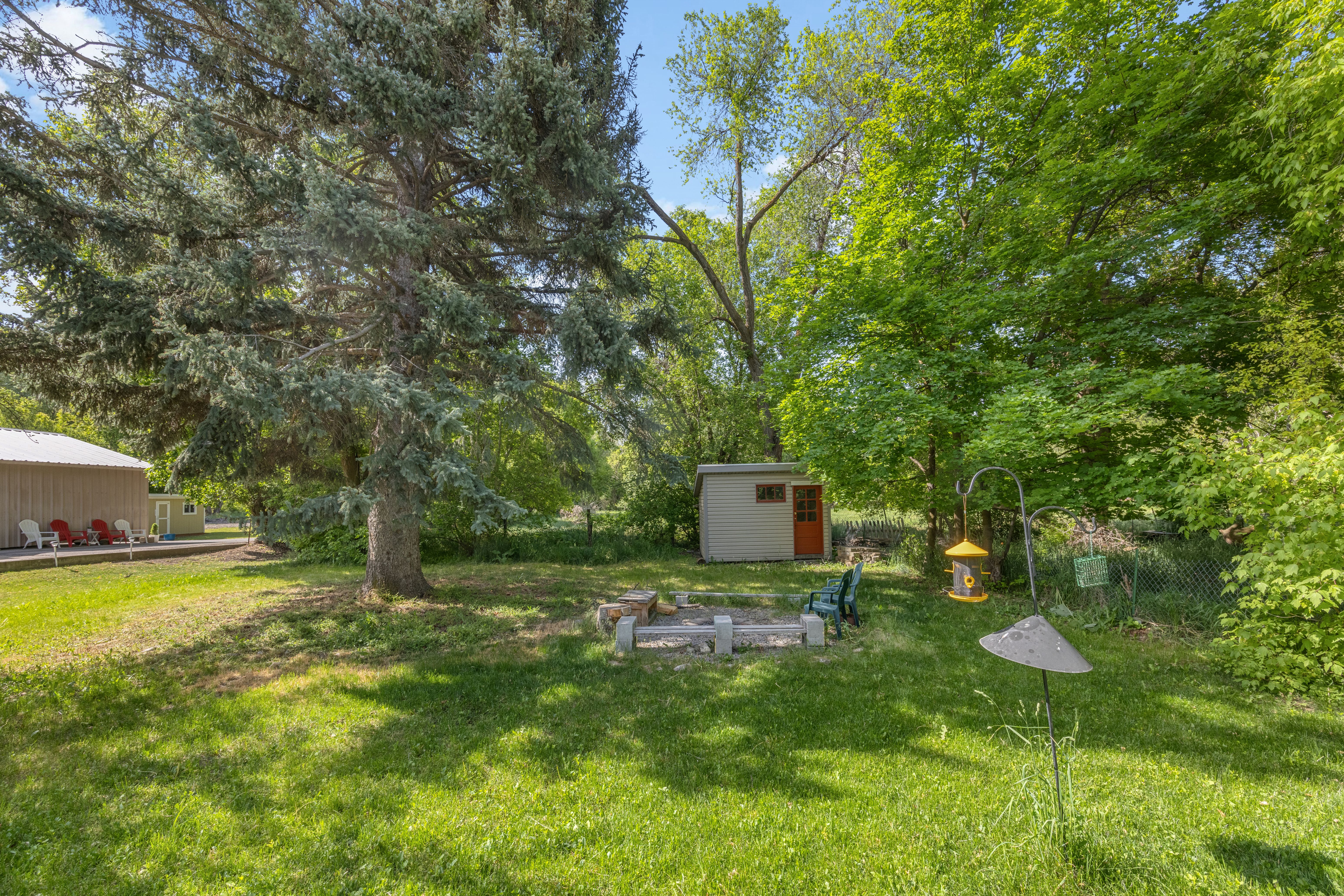Expansive grassy yard featuring mature trees, a built-in fire pit, and a detached storage shed with an orange door
